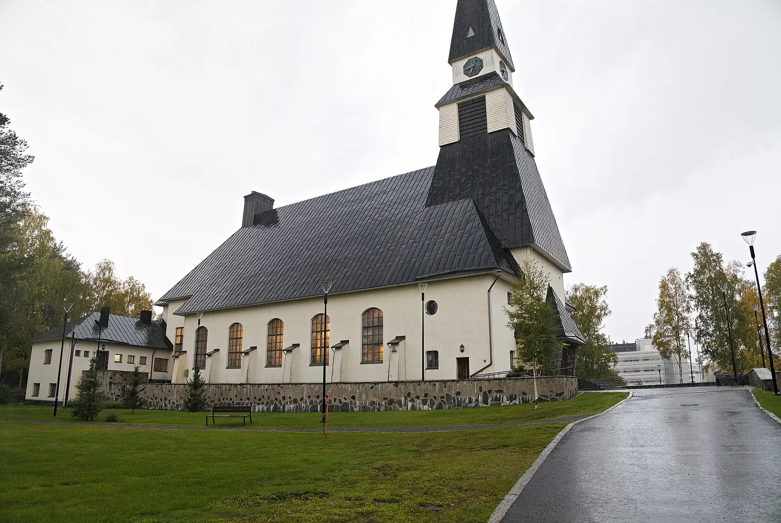Valkoinen kirkko tummalla kattolla ja kellotornilla sateisena syyspäivänä nurmikon takana